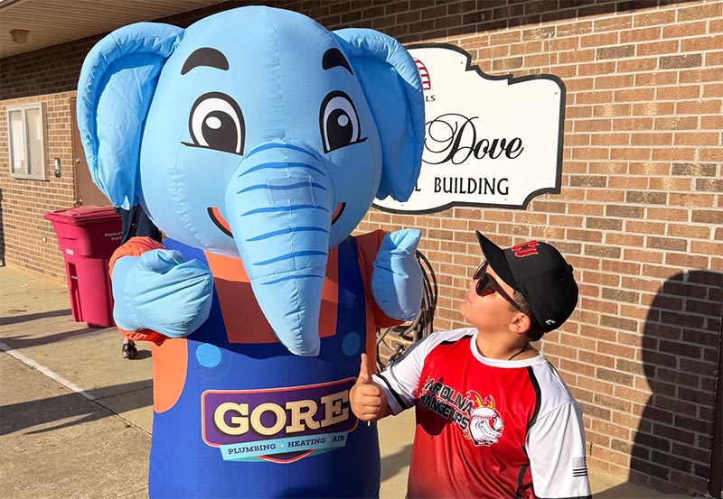Child giving a thumbs-up next to a blue inflatable elephant mascot promoting Gore Plumbing, Heating, and Air outside a building.