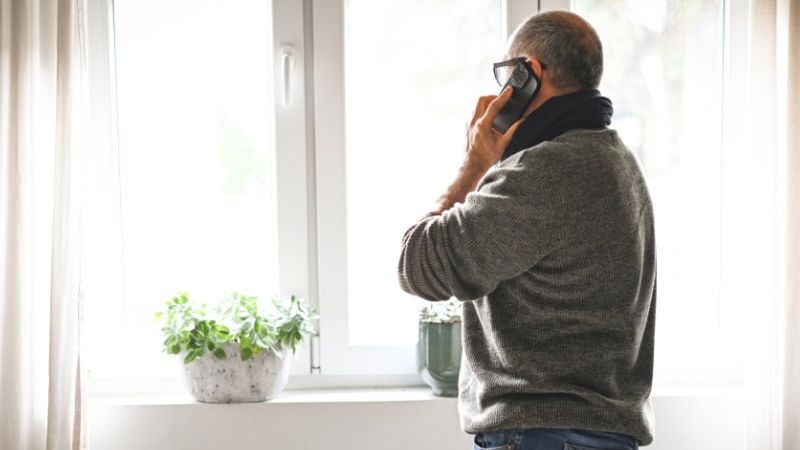 Man on phone by window, wearing glasses and sweater, looking outside. Houseplant on windowsill, bright natural light.