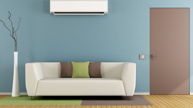 Modern living room with sofa, blue wall, air conditioner, and stylish door. Cozy decor with green and brown cushions.