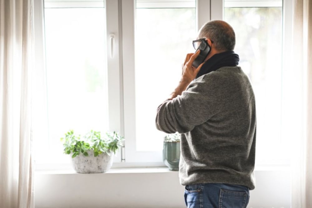 Man on phone by window, wearing glasses and sweater, looking outside. Houseplant on windowsill, bright natural light.
