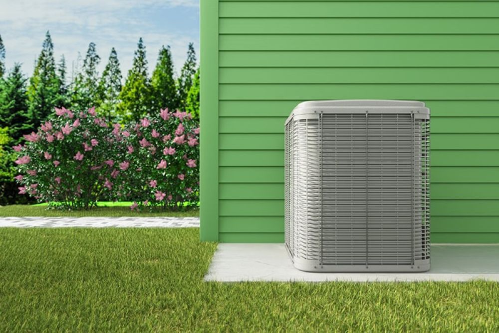 Exterior air conditioning unit next to a green house, surrounded by grass and shrubbery.