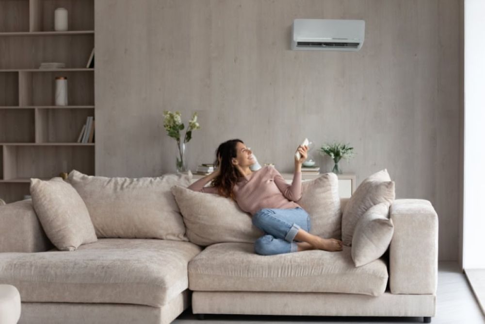 Woman relaxing on a beige sofa in a modern living room, using remote to control air conditioner on the wall.