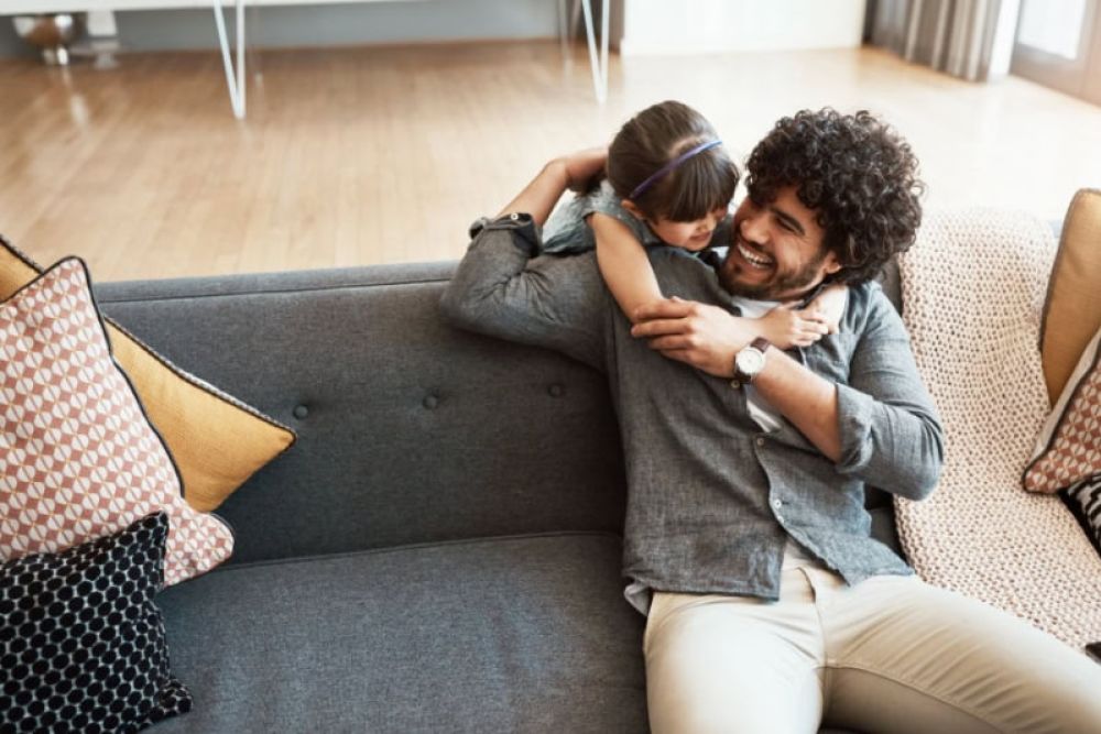 Father and daughter laughing and hugging on a sofa in a cozy living room setting, surrounded by cushions.