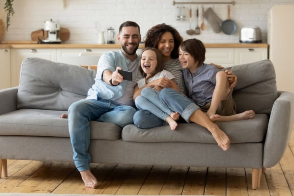 Happy family sitting on a cozy sofa, smiling and taking a selfie together in a bright, modern living room.