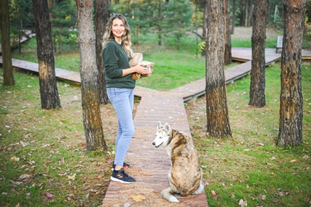 Woman holding basket in forest with dog sitting on path, surrounded by trees and autumn leaves.