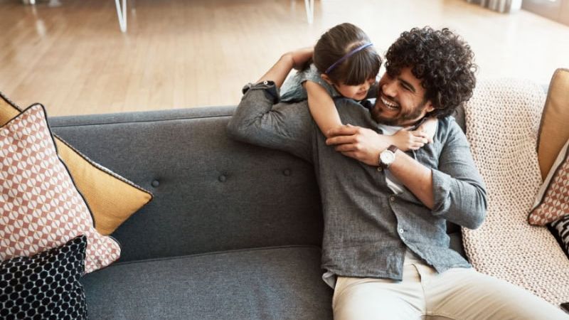 Father and daughter laughing and hugging on a sofa in a cozy living room setting, surrounded by cushions.