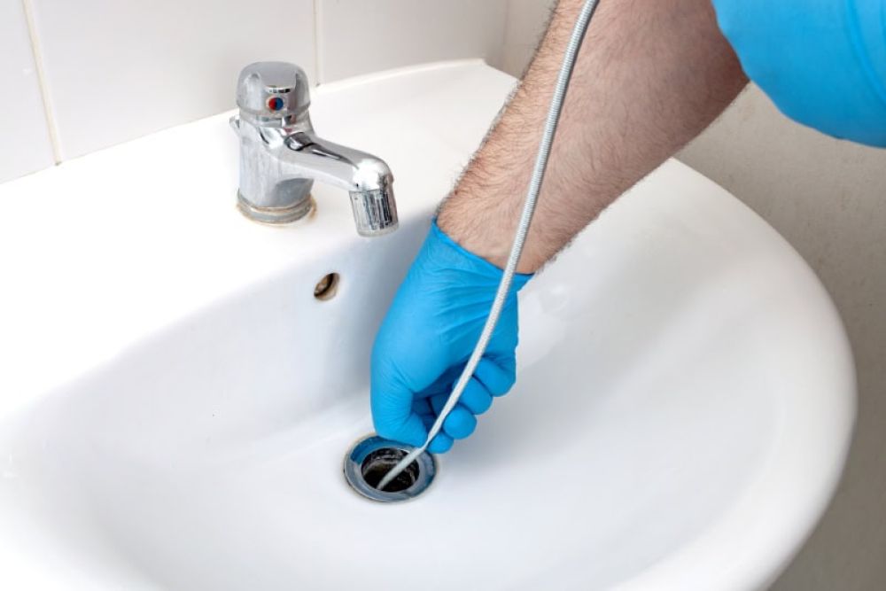 Plumber using a drain snake to unclog a bathroom sink with blue gloves.
