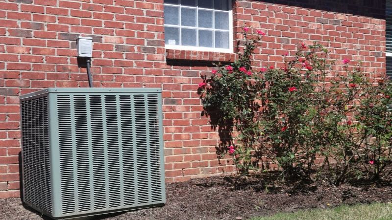 Outdoor air conditioning unit next to a brick wall and rose bushes in a sunny garden setting.