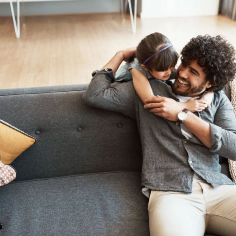 Father and daughter laughing and hugging on a sofa in a cozy living room setting, surrounded by cushions.