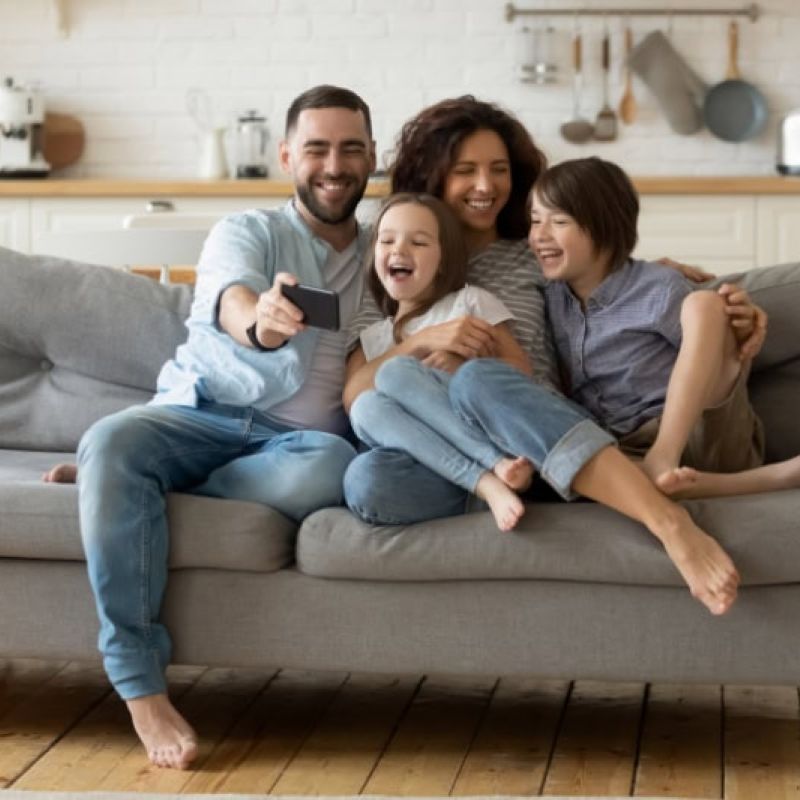 Happy family sitting on a cozy sofa, smiling and taking a selfie together in a bright, modern living room.