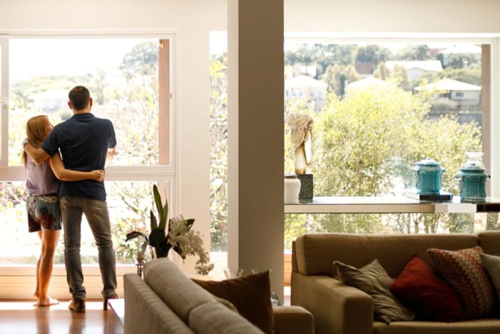 Couple embracing by large window, enjoying serene view, with cozy living room setting in foreground.
