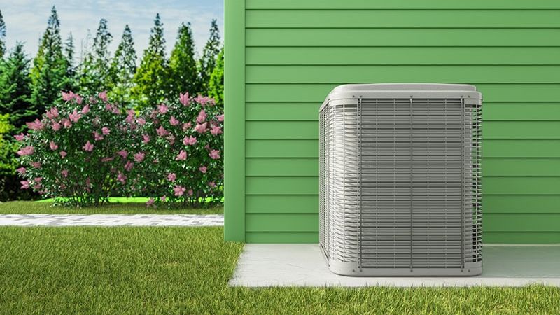 Exterior air conditioning unit next to a green house, surrounded by grass and shrubbery.