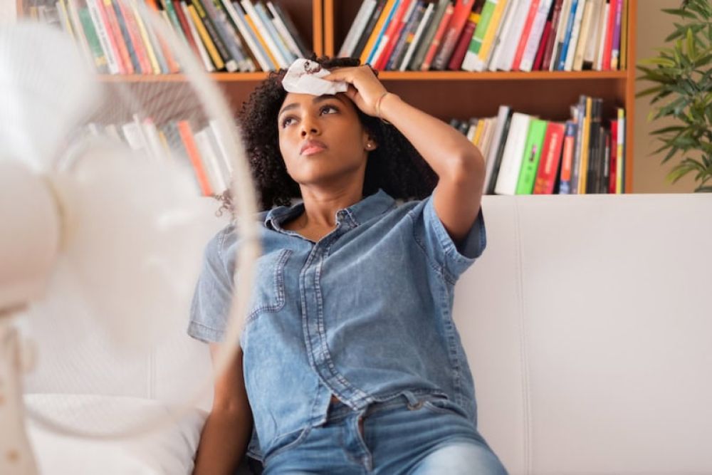 Person cooling off with fan in living room, wiping forehead, sitting on couch, bookshelf in background.