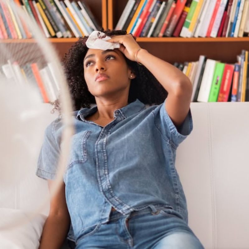 Person cooling off with fan in living room, wiping forehead, sitting on couch, bookshelf in background.