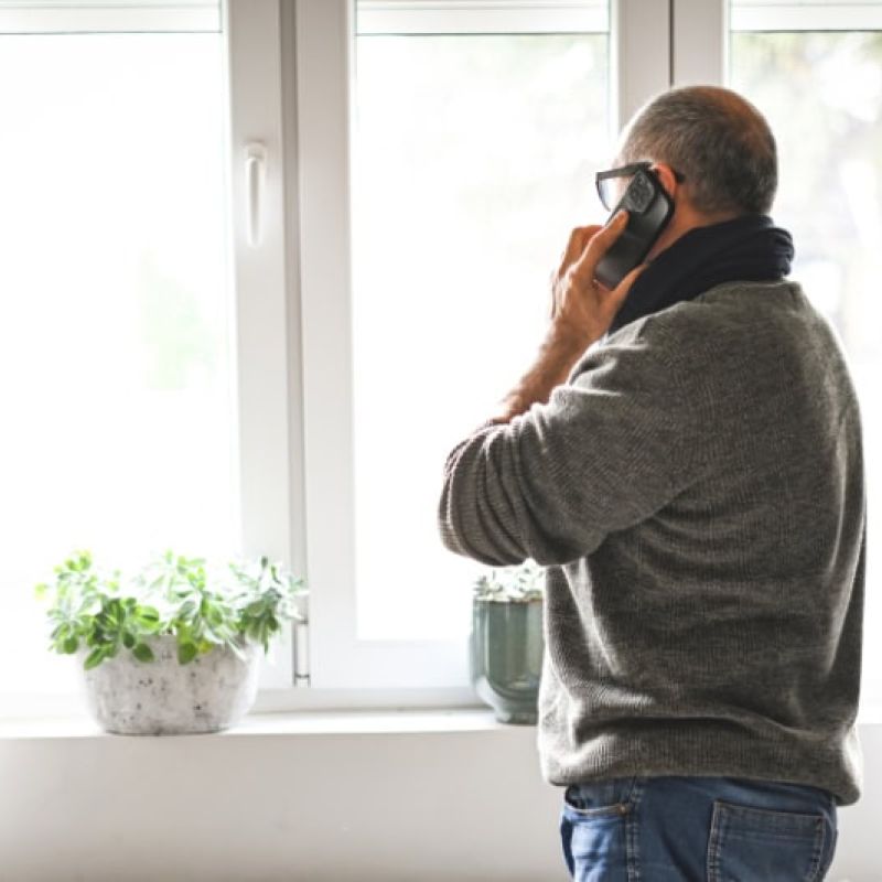 Man on phone by window, wearing glasses and sweater, looking outside. Houseplant on windowsill, bright natural light.