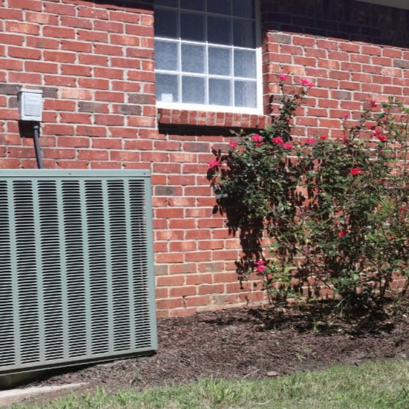 Outdoor air conditioning unit next to a brick wall and rose bushes in a sunny garden setting.