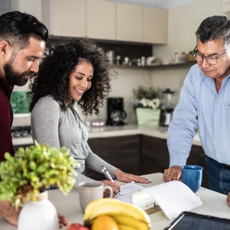 Three people reviewing documents in a modern kitchen, discussing ideas over coffee.