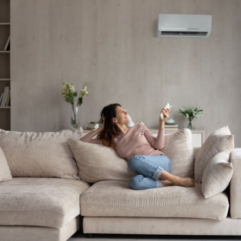 Woman relaxing on a beige sofa in a modern living room, using remote to control air conditioner on the wall.