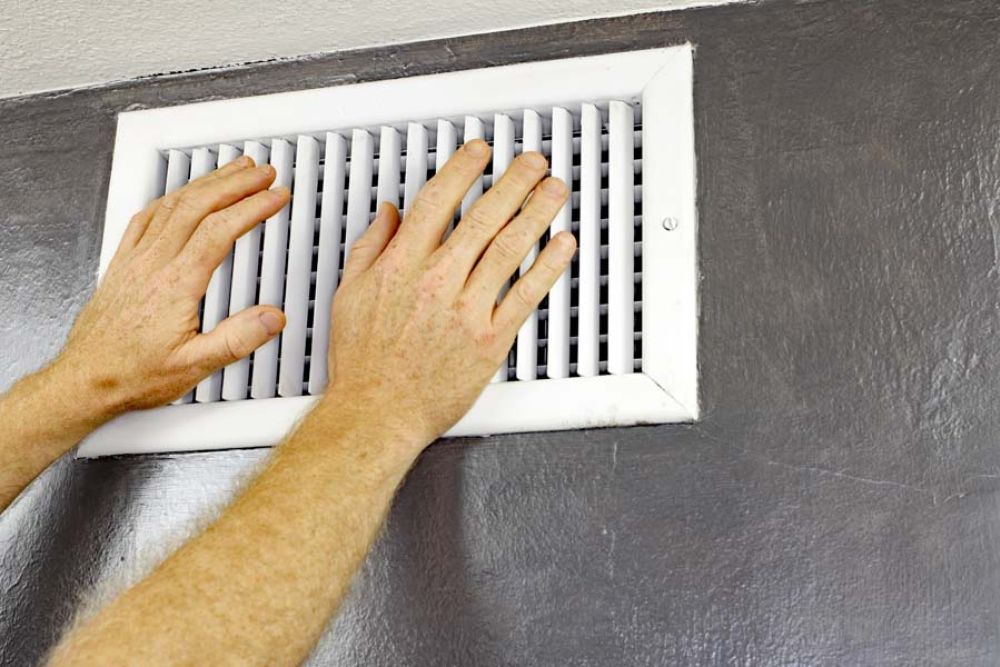 Hands adjusting a white air vent on a gray wall for improved airflow in a home or office environment.