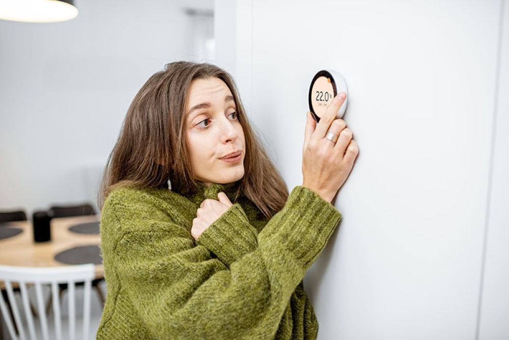 Woman in green sweater adjusts smart thermostat on wall for temperature control.