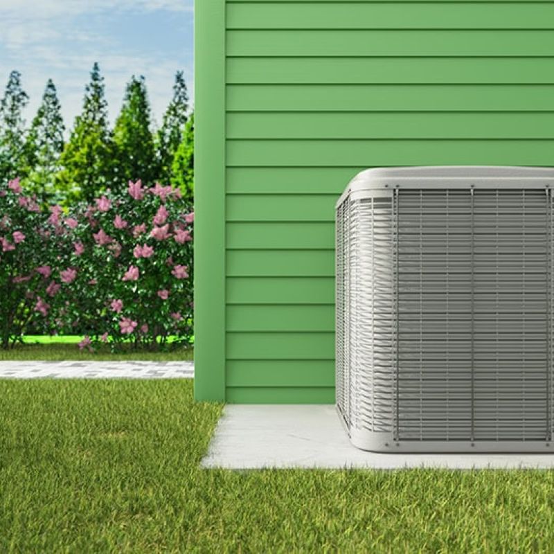 Exterior air conditioning unit next to a green house, surrounded by grass and shrubbery.