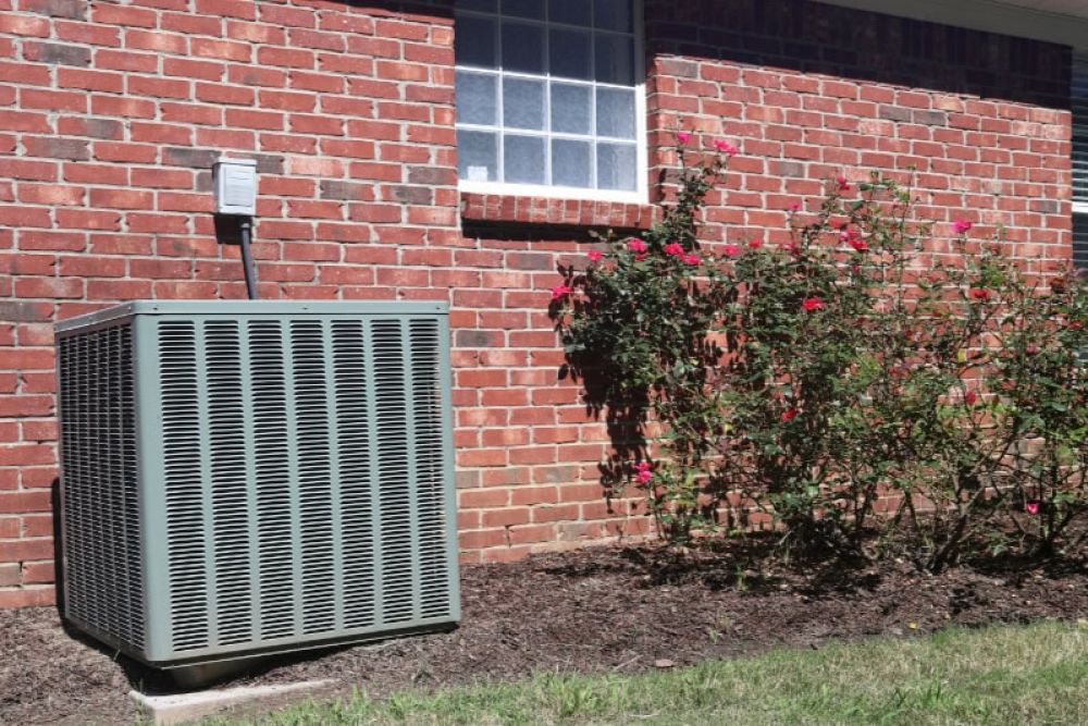 Outdoor air conditioning unit next to a brick wall and rose bushes in a sunny garden setting.