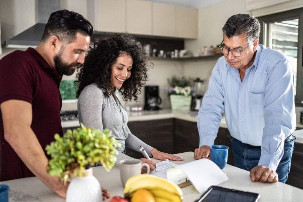 Three people reviewing documents in a modern kitchen, discussing ideas over coffee.