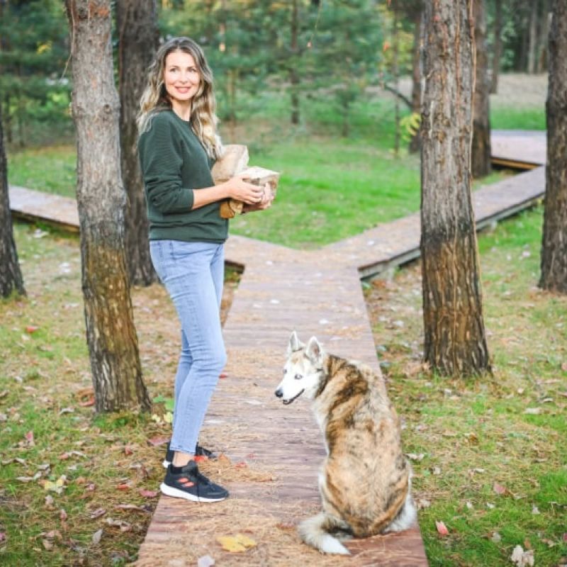 Woman holding basket in forest with dog sitting on path, surrounded by trees and autumn leaves.