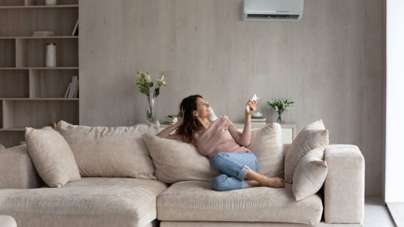 Woman relaxing on a beige sofa in a modern living room, using remote to control air conditioner on the wall.